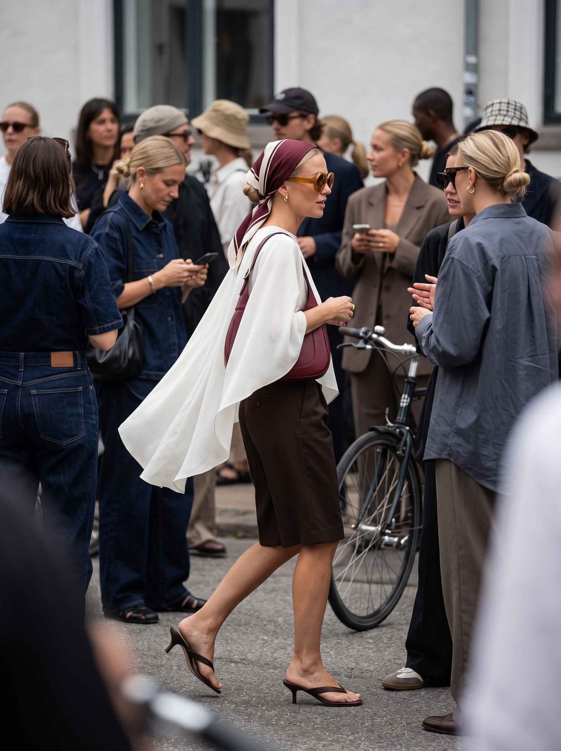 Copenhagen fashion week 2026. Kvinna på gatan klädd i ljus cape top, bermuda shorts, flip flop kitten heels, retro solglasögon, siden scarf och väska i vinröda toner.