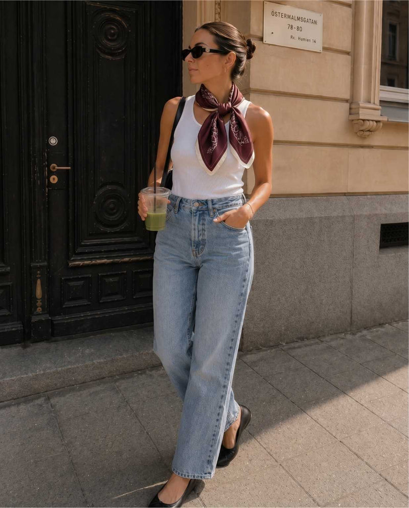 Kvinna i avslappnad elegant outfit, klädd i vitt linne, mörk röd blommig sidensjal och blå jeans
